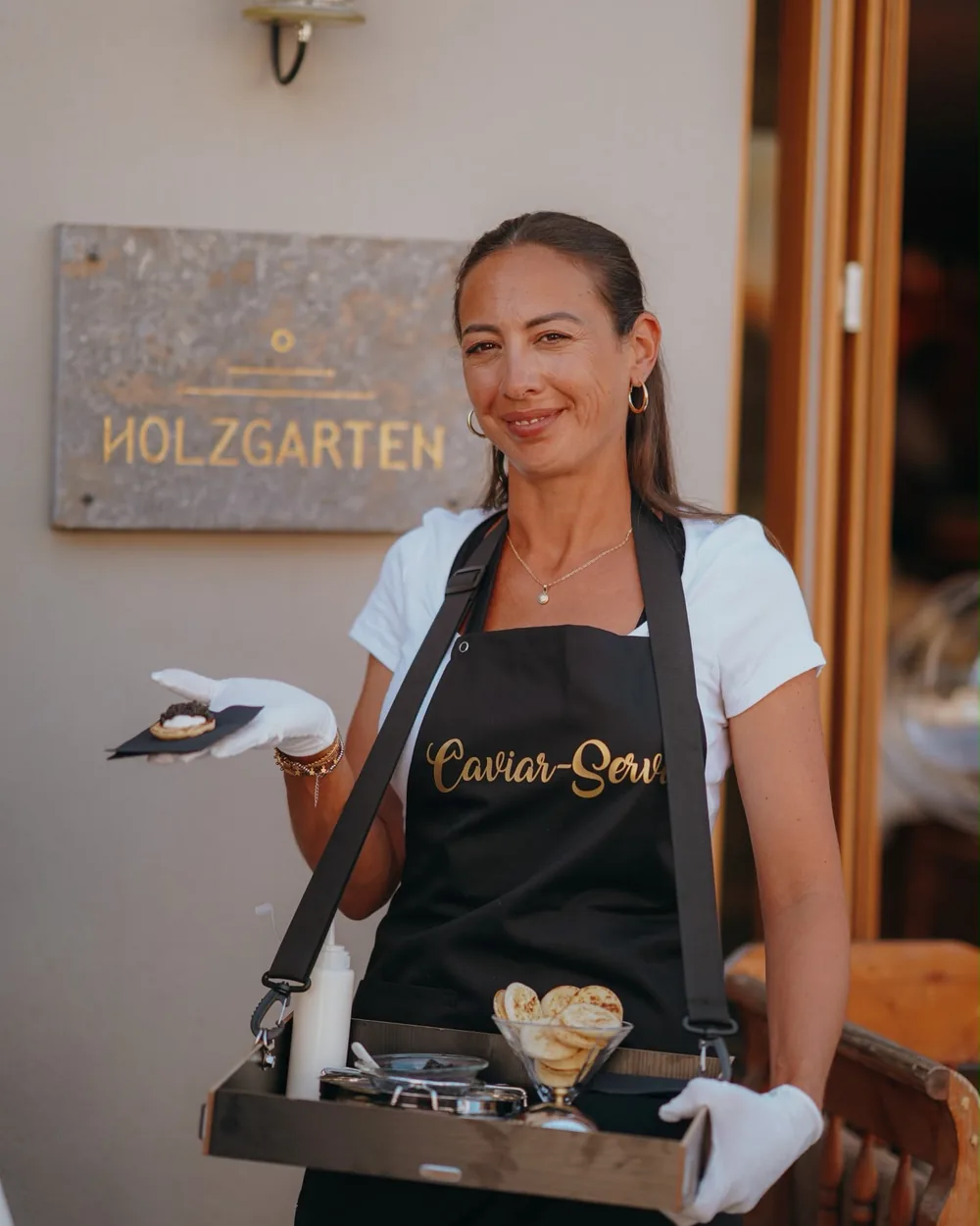 Servicekraft mit Caviar-Service-Schürze lächelt mit einem Tablett voller Blinis in die Kamera