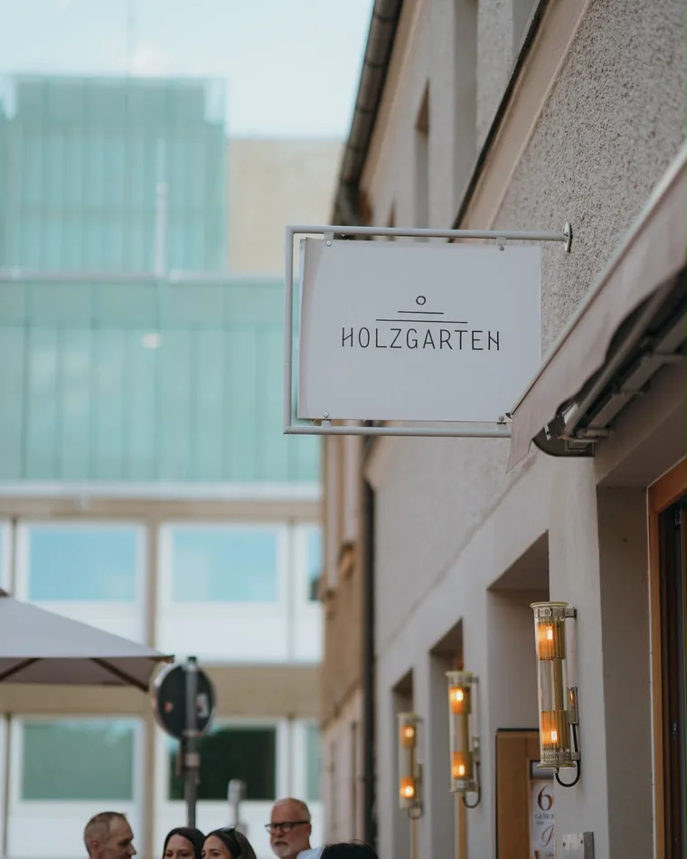 Schild mit der Aufschrift Holzgarten an einer Hauswand, darunter Gäste in Weiß mit Sektgläsern
