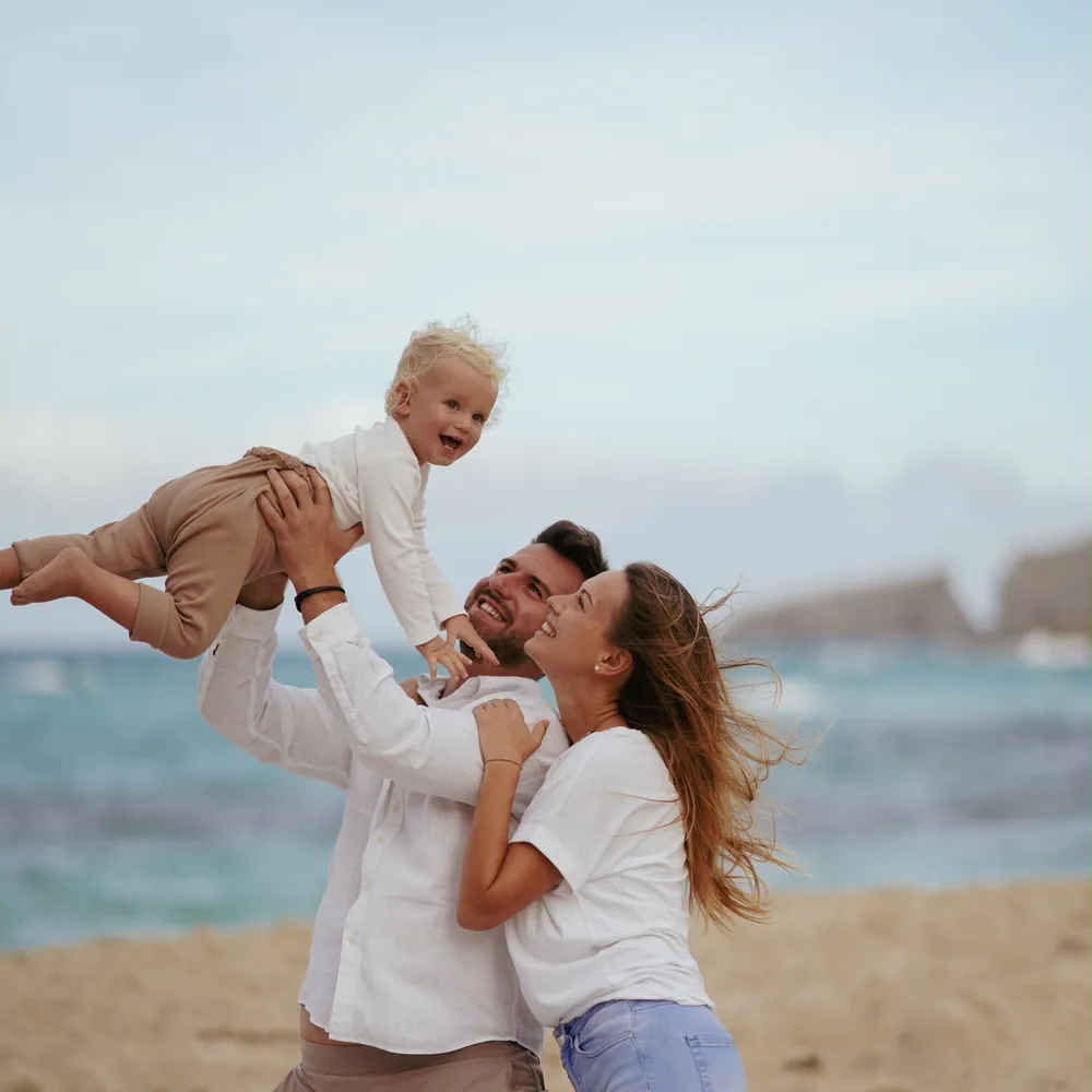 Vater hebt sein lachendes Kleinkind am Strand hoch, die Mutter lacht daneben