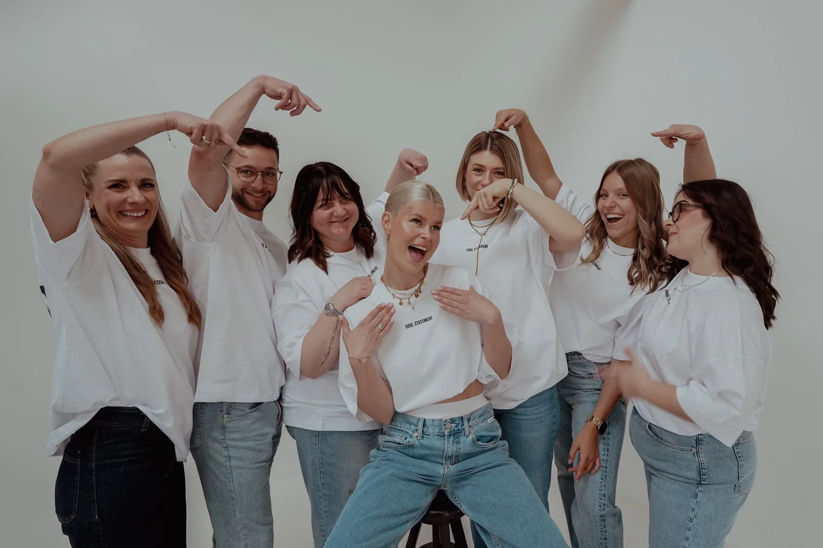 Siebenköpfiges Team in weißen Shirts im Studio, alle zeigen lachend auf die Frau in der Mitte