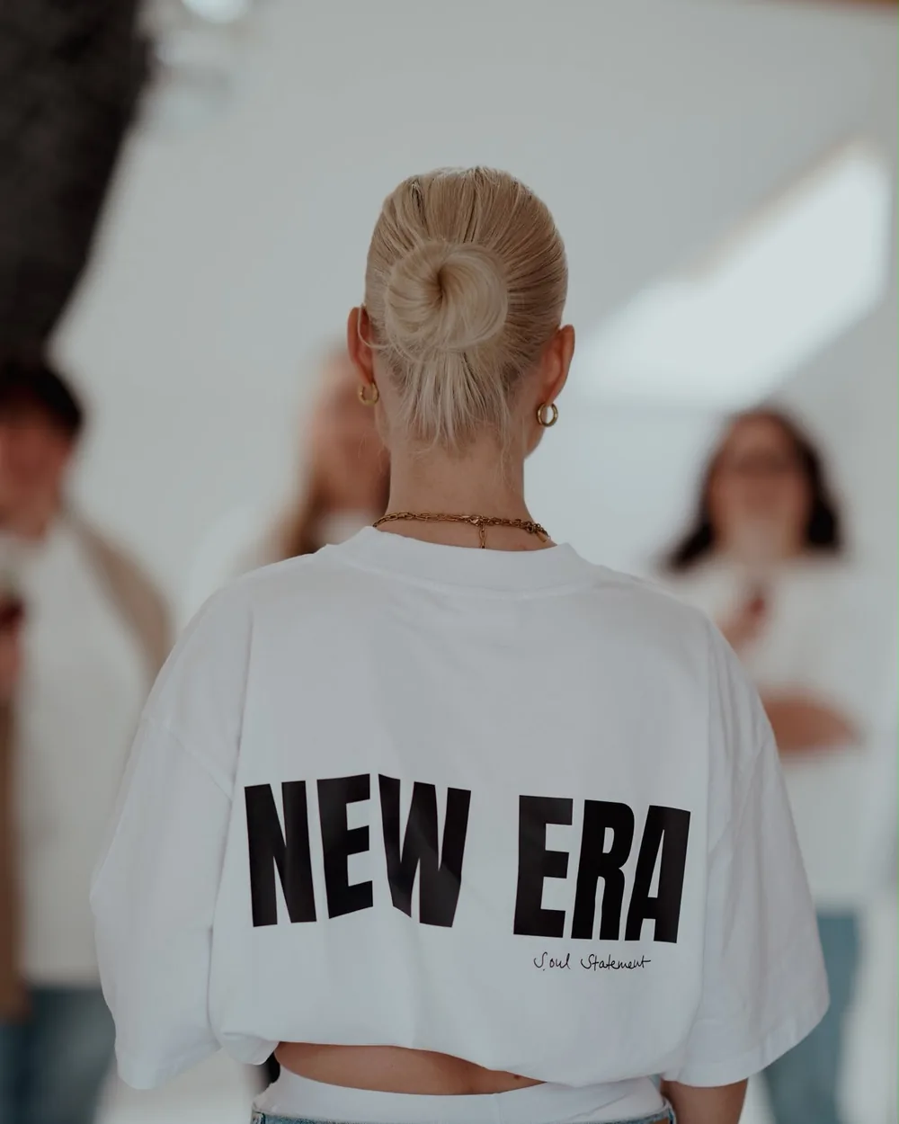 Hinter den Kulissen eines Shootings: Frau im weißen Shirt mit dem Aufdruck New Era, dahinter unscharf das Team im Raum
