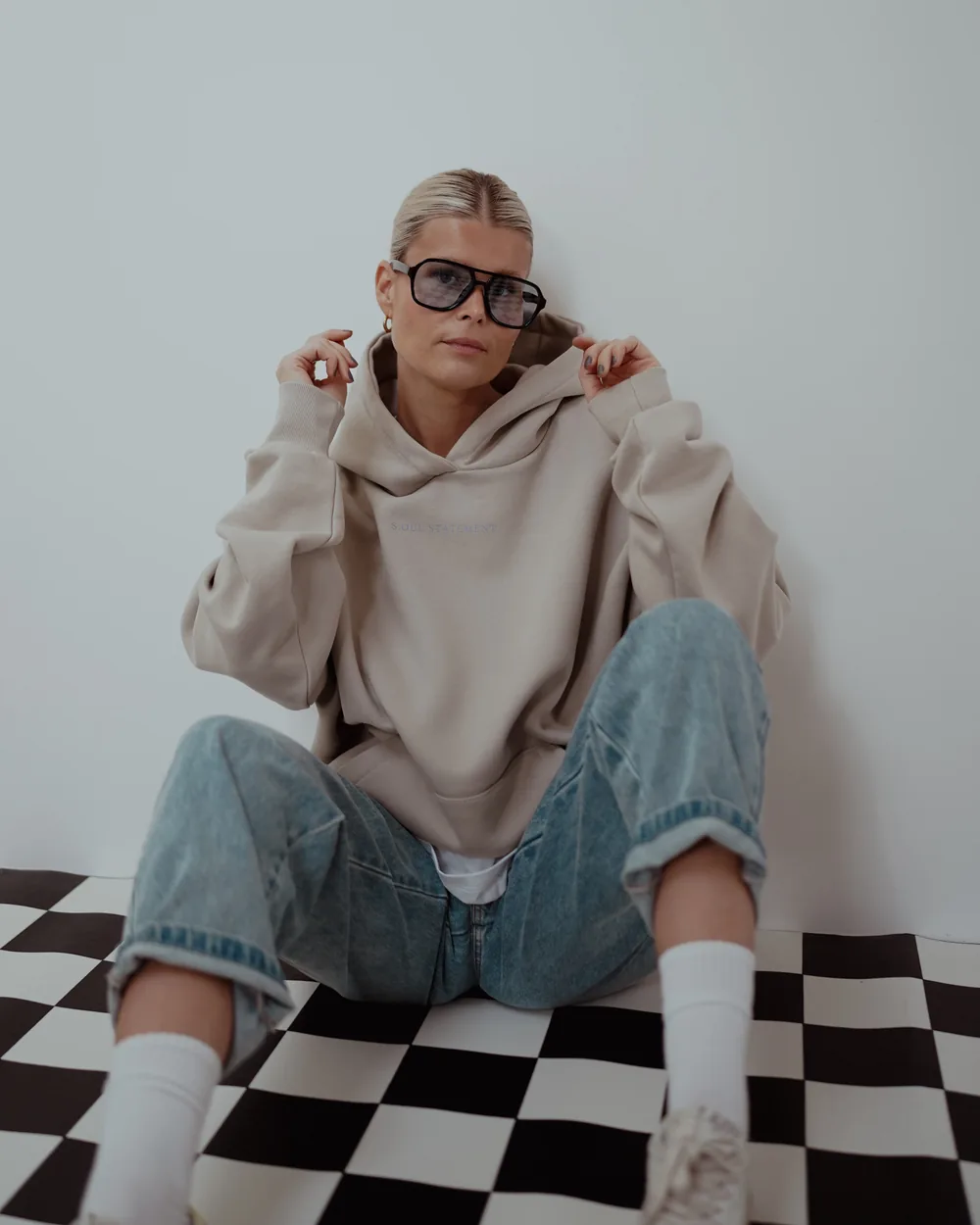 Markenshooting im Studio: Frau im beigen Hoodie sitzt vor einer weißen Wand auf einem Schachbrettboden