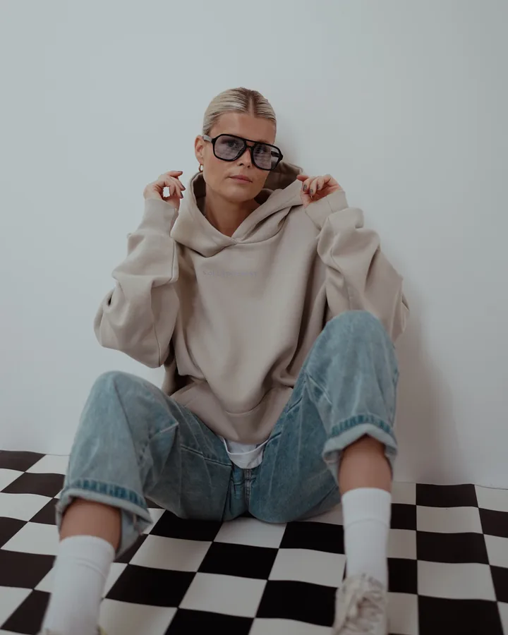 Frau im beigen Hoodie mit Sonnenbrille sitzt vor einer weißen Wand auf einem schwarz-weißen Schachbrettboden